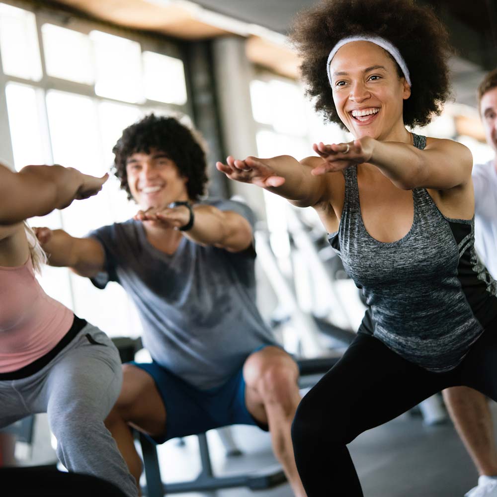 Group of people smiling and performing squats together in a brightly lit gym during a fitness class.