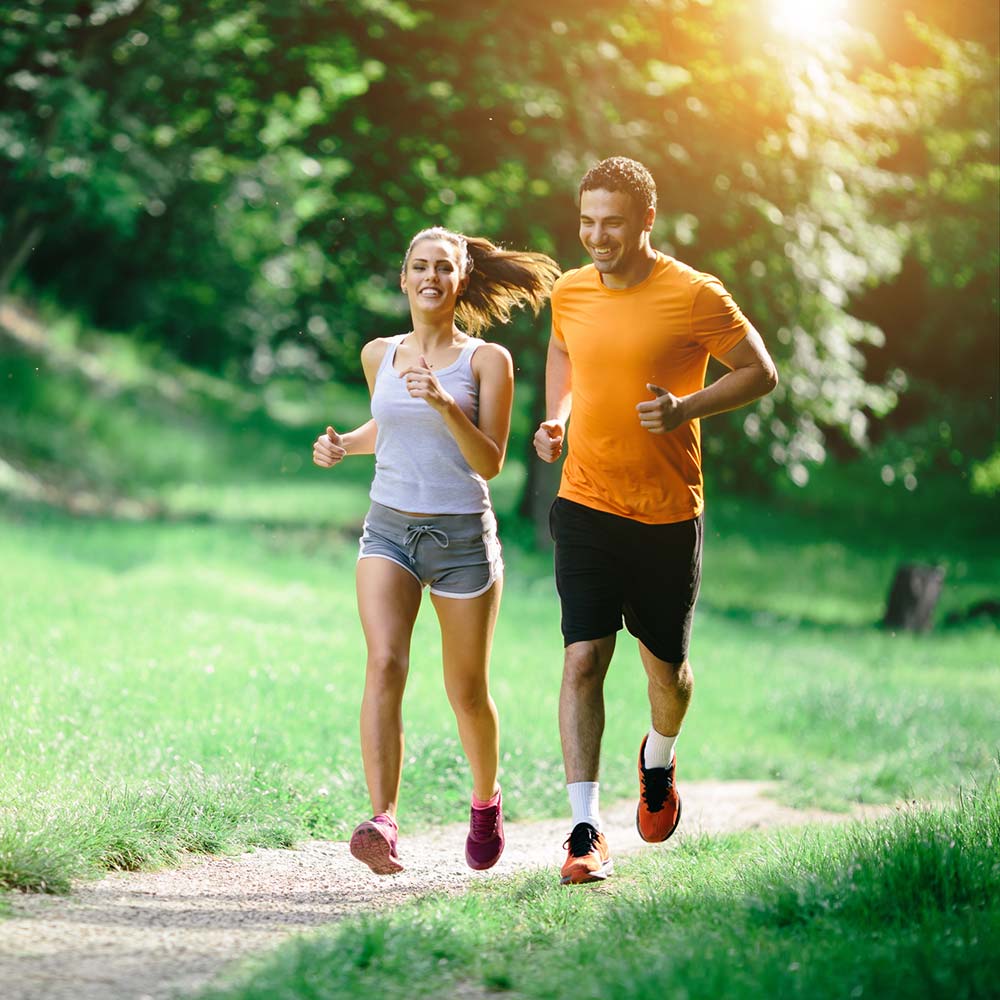 A smiling man and woman jog together on a sunlit park trail surrounded by lush green trees and grass.