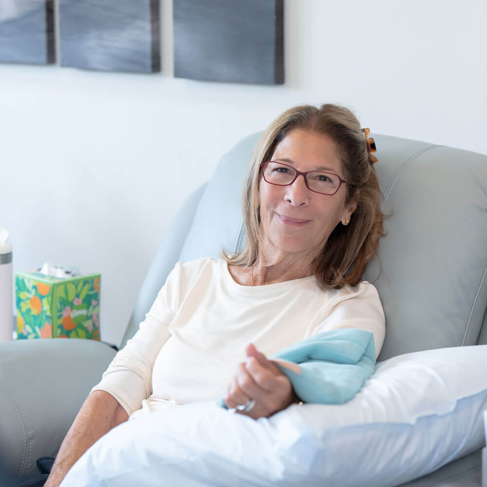Smiling woman seated in a recliner during IV therapy with a pillow supporting her arm.